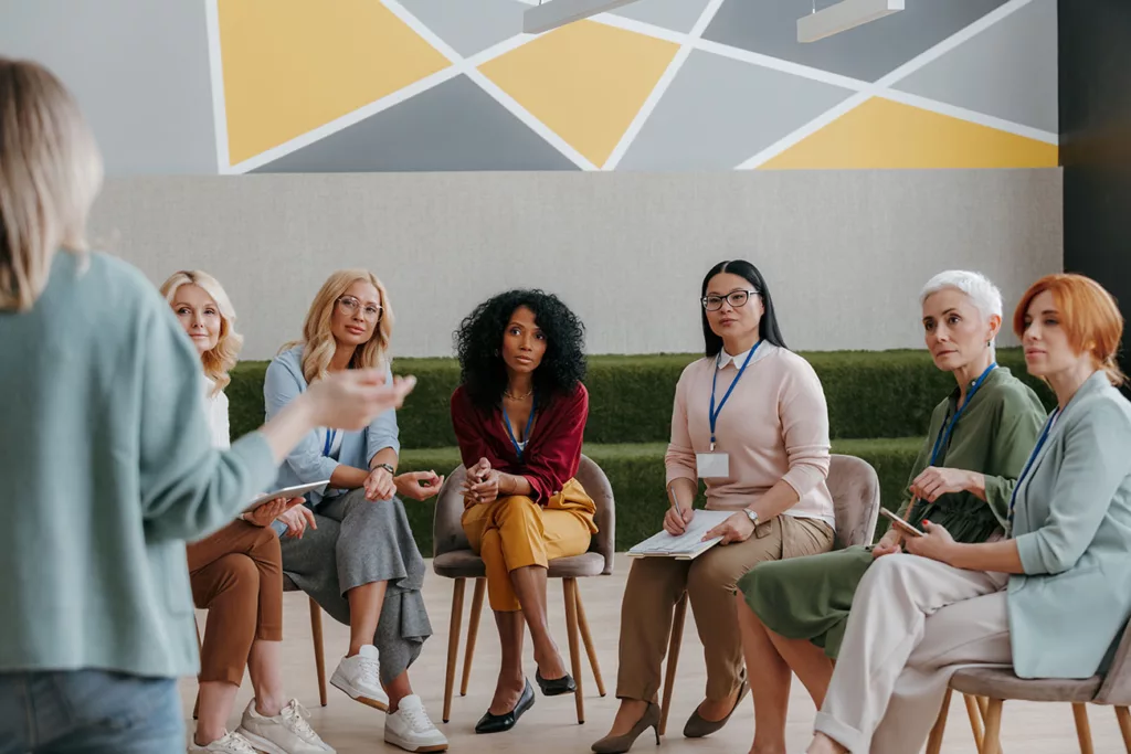 Multi-ethnic group of women in casual to casual business attire listening to a speaker while sitting in a semicircle of chairs. 